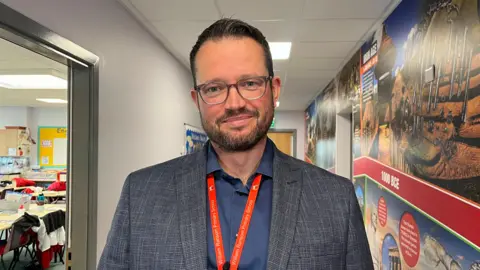 Nicola Haseler/BBC A man smiling in a school corridor. He is wearing glasses, a grey blazer and a navy shirt. He has a red lanyard with William Ransom Primary School written on it.