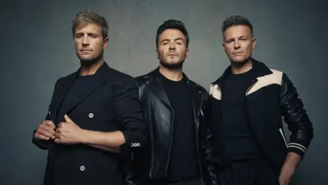 Westlife The picture features three men wearing dark jackets and standing beside each other. The three men have dark t-shirts on and are standing in front of a grey background.