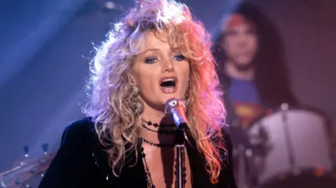 Bonnie Tyler singing into a microphone. She has big blonde hair and a black dress on. Behind is a drummer - he is slightly blurred, as Bonnie is the focus of the image.