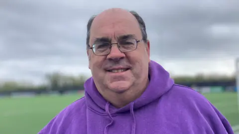 A man with glasses and a purple hoody is pictured smiling at the camera in front of a football pitch.