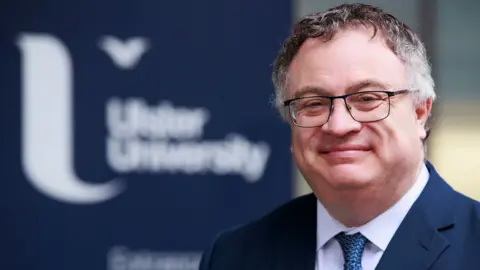 A man with grey and dark brown hair smiles in the direction of the camera. He is wearing dark coloured metal rimmed glasses, a navy blue suit jacket, a white shirt and a blue patterned tie. The background is out of focus, but you can make out the navy blue and white Ulster University branding.