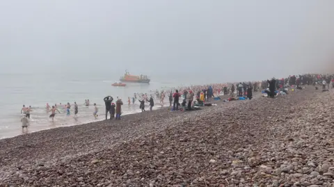 Dozens and dozens of people about to part in a Christmas swim at Budleigh Salterton with some already in the water. There is a large crowd of onlookers watching the swim. It is a misty day and two RNLI boats - one large and one small - can be seen on the water.