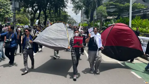 A group of protesters walk down the street in central Jakarta. Several of them are holding banners and posters that protest against the revisions to Indonesia's military law. In the foreground are a few protesters holding a grey tent and a maroon-coloured tent. 