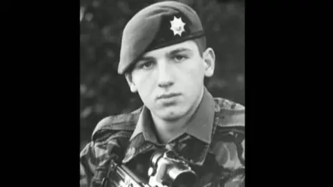 Supplied Lance Corporal Alan Forcer in his military uniform. He is wearing a beret with multi-pointed star badge and has on Army camouflage. The sight of the weapon he is holding is just visible in the bottom of the black and white picture.