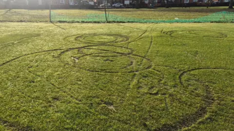 The tracks left behind by vehicles used on a cricket ground, they are large indentations to the grass. They are in circular patterns in various places, beyond them is green plastic fencing and cars and houses in the background.