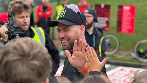 Sir Mark Cavendish, wearing a black cap with sun glasses on top of it, giving a high five to a young fan while a man with a film camera looks on.