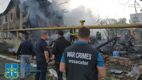 Odesa Regional Prosecutors Odesa Regional Prosecutors stand at the site of a Russian drone attack on the port city of Izmail. The man closest to the camera has a vest reading "WAR CRIMES prosecutor". They are looking at a house that has been partly destroyed, there are lots of blackened and charred ruins, and it's still smoking heavily. In the background are firefighters hosing down the smoking ruins with water, and there is also a digger-type vehicle off to the side.