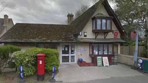 A view of the Post Office in Foxton. The small building has brown bay windows and a red post box outside.