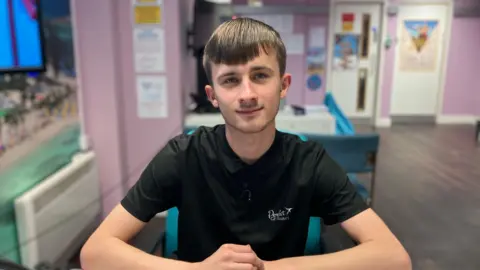 Abi Smitton / BBC News A man with brown hair sits at a desk, wearing a black polo neck shirt.