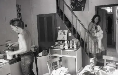 Alan Van Wijgerden The black and white image shows a shirtless man at a kitchen sink, kettle in hand, yawning. A woman is coming down the stairs in the background, and a kitchen table and sideboard can be seen strewn with empty beer cans, and kitchen containers. 