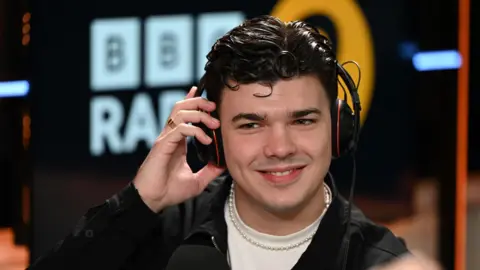 Harry Clark, wearing a pair of headphones in the BBC Radio 2 studio wearing a white t-shirt. a gold chain and a black jacket