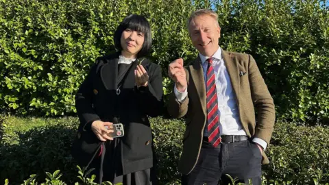 Yuriko Shigyo stands in front of a bush. She is wearing a black coat, white shirt and black jumper. She is holding a leaf in her left hand. Next to her is Jonathon Jones from Tregothnan. He is wearing a tweed blazer, a white shirt and a red and black striped tie. He is holding a leaf in his right hand.