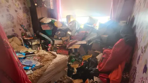 Image shows Ruth Cookson's bedroom when her hoarding was at its worst. Rubbish, boxes, clothes and other items are piled high up the walls and blocking out the light from the window. 