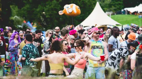 A group of festival-goers in brightly-coloured clothing looking happy and relaxed. Two are dancing in the foreground and in the background there is a large tipi tent.