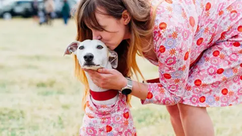 North East Dog Festival A young woman kisses her dog on the forehead. They are both wearing tunics made from the exact same fabric - a chaotic pattern of pastel pink and white flowers.