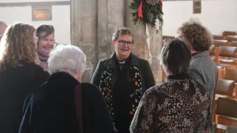 Diocese of Lincoln A group of six women talking, four have their backs to the camera and two are facing the camera. The woman at the centre of the photograph is wearing a dark green coat and a scarf with flowers on it, of different colours, she has grey hair and dark rimmed glasses.