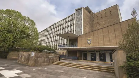 Durham County Council Steps lead to the entrance of Durham County Hall. The council's blue and yellow crest is attached the beige ageing building, which is covered in multiple windows.