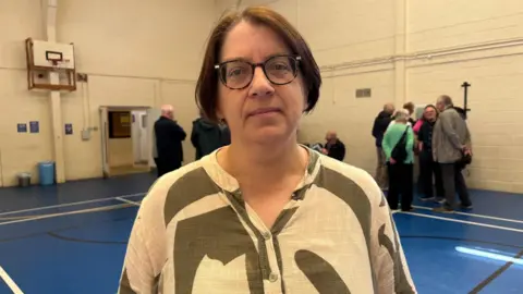 Simon Thake A woman with short dark hair stands in a sports hall. Behind her a group of adults are huddled together talking and looking at maps on the wall