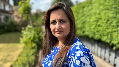 Saj Golby on a sunny day. She is an Asian woman wearing a bright blue and white dress. The background is blurred but we can see some bushes behind her.