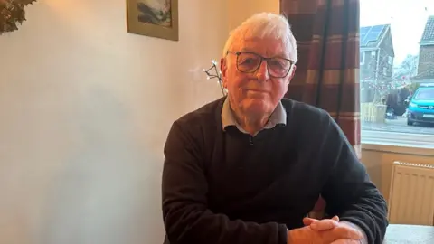 Keith Bell smiling into the camera. He has got white hair and is wearing glasses. He is sitting in front of a window with tartan curtains.