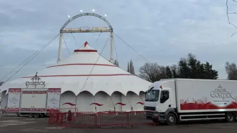 Irina Archer A circus big top, white with red trim. A nearby lorry, also white with some red patterning on the side, is emblazoned with a logo reading "CIRCUS CORTEX".