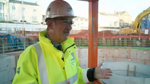 BBC A man in a high-viz jacket and hard hat stands in front of a large concrete water storage tank that is under construction. There is building equipment in the background. 
