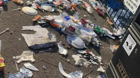 A close-up of overflowing rubbish from a bin strewn across a promenade, next to a black bin and black railings.