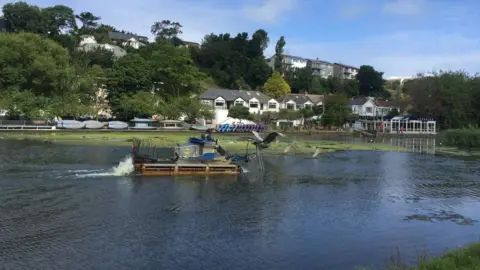 The boating lake with clear water. There is a small dredging boat moving across it. Behind the lake is a row of white houses, tress and some small watercraft stored upside down.