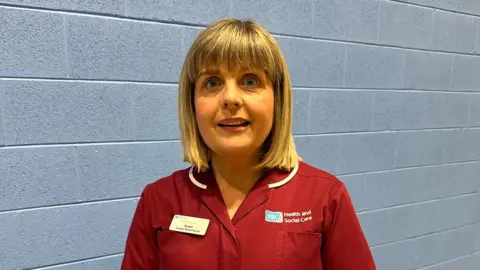 Susan is standing in front of a grey brick wall. She has short blond hair and is wearing a red medical top with a name badge on one side and 'Health and Social Care' on the other.