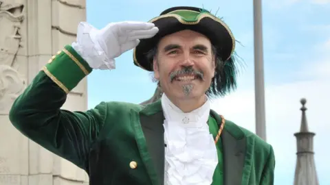 Michael Wood - a man with a dark, greying moustache and goatee beard - salutes while wearing a green and black town crier's uniform.