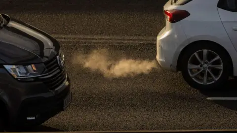 Two cars, one black and one white, driving down a road. There are fumes coming out of the exhaust of the white car.