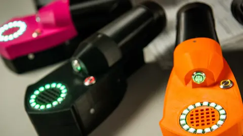 A close-up of three of the portable testing devices. They have a round tube which leads to a section topped with a dial of tiny lights and a ventilation grid. 