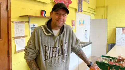 A photo of a man stood over crates of food at the Phoenix centre in Matson. He is wearing a grey hoodie and a dark cap and is looking at the camera