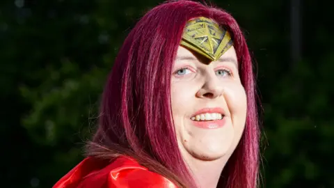 Nick Butcher Photography/St Elizabeth Hospice A woman with long red hair and a superhero headpiece smiles at the camera. She also dons a red superhero cape. 