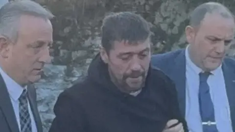 RTÉ Joe Butler, being escorted by two men in suits who are holding him by the arms. He is wearing a dark coloured hooded jumper and behind him is a stone wall. 