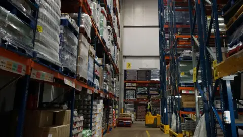 A warehouse with shelves of stock. There is a concrete floor with yellow markings.