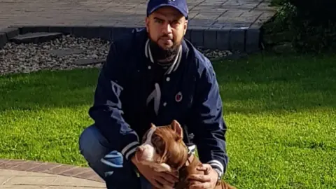 Moheiz Adam crouched by a tan and white dog, which appears to have cropped ears. Adam is wearing a blue baseball hat and navy jacket and jeans, and has a dark, short beard. 