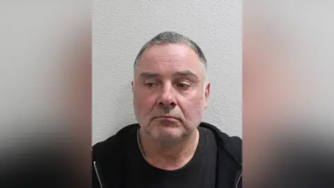 Custody image of Thomas McKenna, wearing a dark top, photographed against a plain wall.