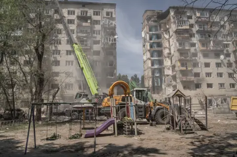 A cruise missile destroyed an entire section of a nine-storey residential building in Kyiv. Flowers were laid by mourners in the playground beneath.