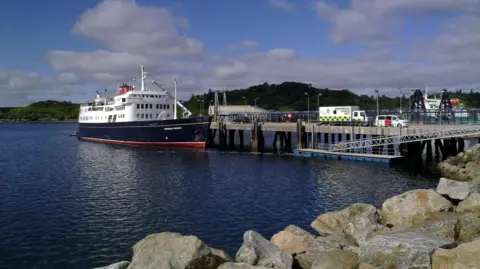 A blue and white hulled cruise shop moored to a jetty with islands behind and rocks in the foreground.