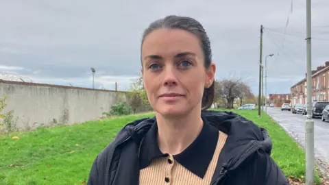 Councillor Lucy Williams stands outside on a roadside in front of a patch of grass. She is wearing a black coat and has brown hair pulled back into a ponytail. 