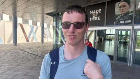 Stephen. He has short dark hair, wearing sunglasses, a light blue t-shirt and carrying a backpack.