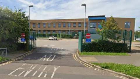 The image shows the entrance to a car park in front of a school. The building has a light brown exterior with rectangular windows in a row along the upper floor. In the foreground, the road surface has large white painted text reading no entry. On both sides of the entrance, there are red no entry signs mounted on green metal gates or posts. 