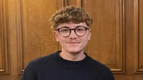 Leo Cavanagh A young man in black-framed glasses with curly fair hair stands in a wood panelled room, smiling. He is wearing a black jumper.
