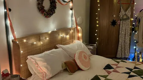 IWC Media Bedroom with a wicker headboard decorated with fairy lights, colourful pillows, a patchwork quilt. A sparkly dress is hanging on the wardrobe which is also draped in fairy lights. There is a garland on the wall.