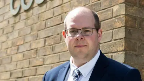 Jonathan Ash-Edwards in a suit and tie outside a court building. He is partially bald and is wearing glasses.