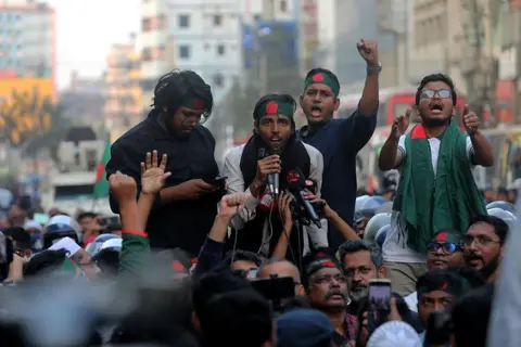 Getty Images Polisi memblokir program ''Pawai ke Komisi Tinggi India'' di Dhaka, Bangladesh, pada 17 Desember 2025. Program ini diselenggarakan untuk menuntut pemulangan mantan Perdana Menteri Sheikh Hasina, yang digulingkan dari kekuasaan menyusul pemberontakan mahasiswa dan masyarakat setelah pembantaian bulan Juli, dan orang lain yang diduga terlibat dalam pembunuhan yang saat ini tinggal di India. Protes ini juga mengutuk konspirasi yang sedang berlangsung oleh proksi India, partai politik, media, dan pejabat pemerintah. (Foto oleh Maruf Rahman/NurPhoto via Getty Images)