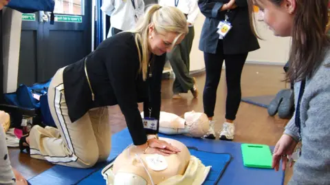 A woman, with long blonde hair, tied back, wearing a black top and cream trousers, leaning over a dummy trying out resuscitation skills. There is another woman wearing and grey top to the right and other people standing around, but you can only see their legs. They are in a school room with a double door to the left. 