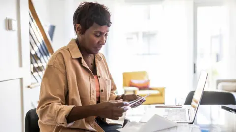 A woman looks at a an envelope with bills while sitting in front of a laptop