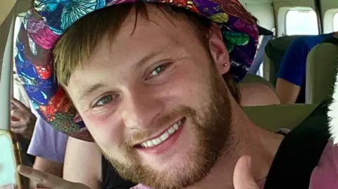 Sam tilts his head and smiles for the camera while sitting as a passenger in a people carrier with others behind him. He is wearing a multicoloured hat